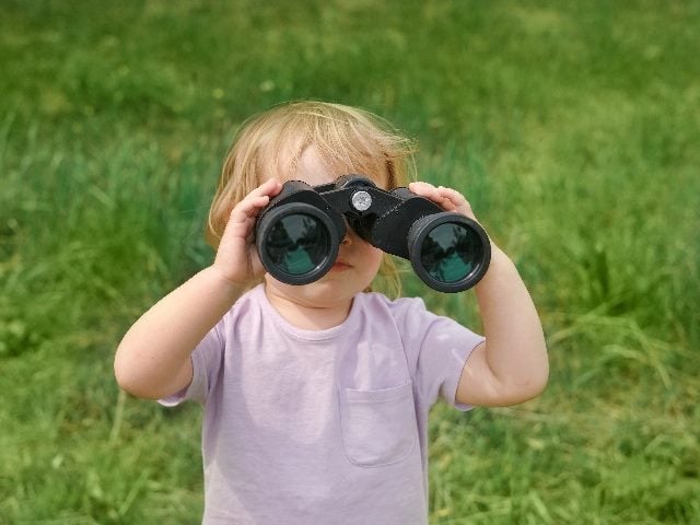 A child with blond hair looks through a pair of binoculars with green grass in the background in piece about digital customer experience trends 2024. 