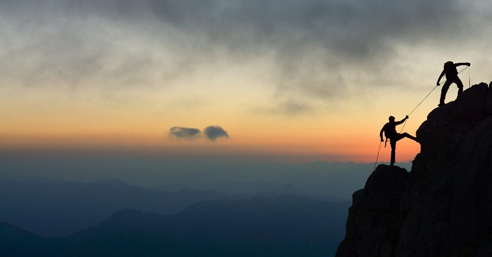 One mountain climber pulls up another mountain climber with a rope with a dramatic sunset in the background in piece about B2B Demand Generation Strategies.