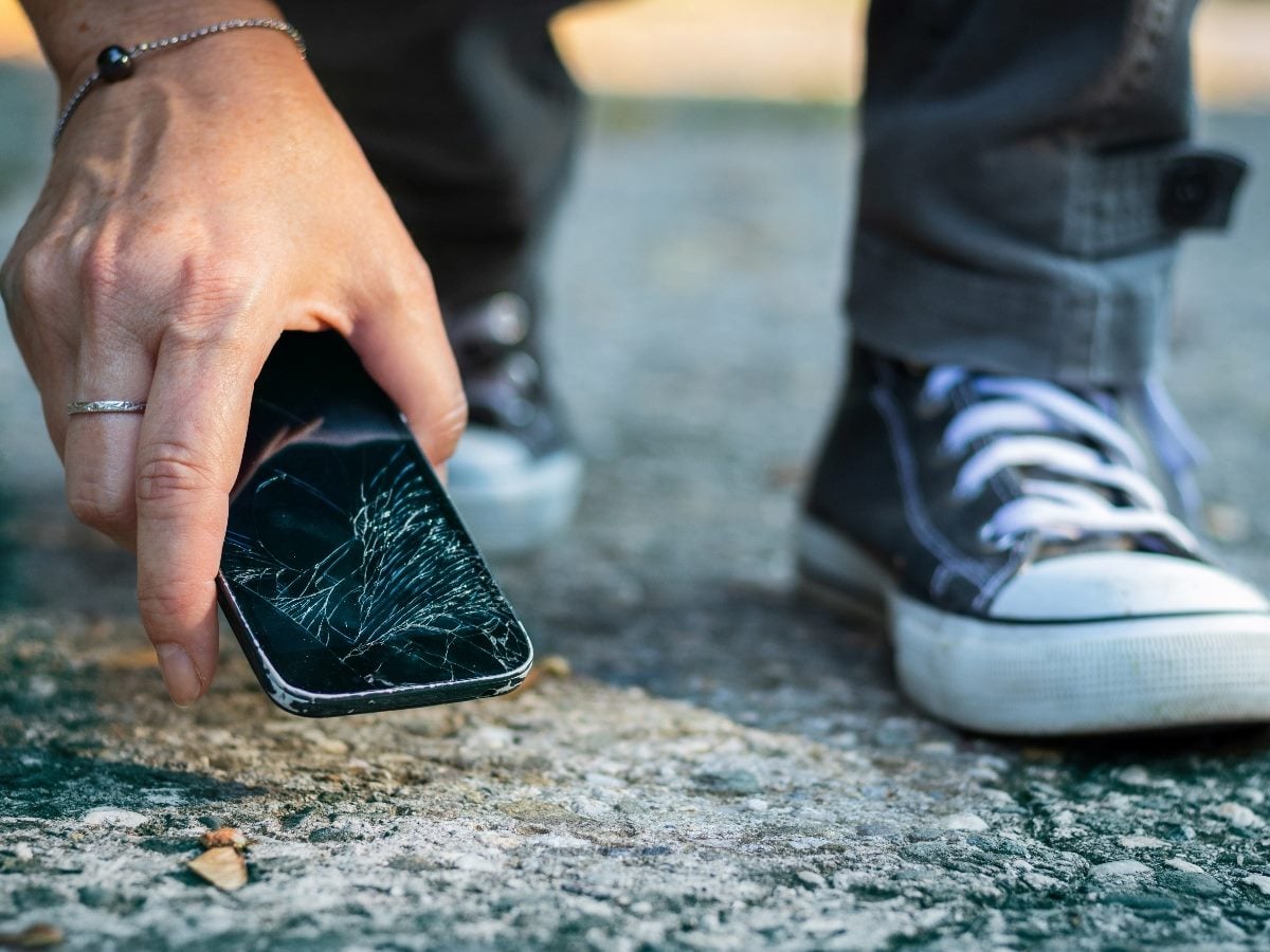 Close-up of a person picking up a smartphone with a severely cracked screen from rough pavement, with casual sneakers visible in the background.
