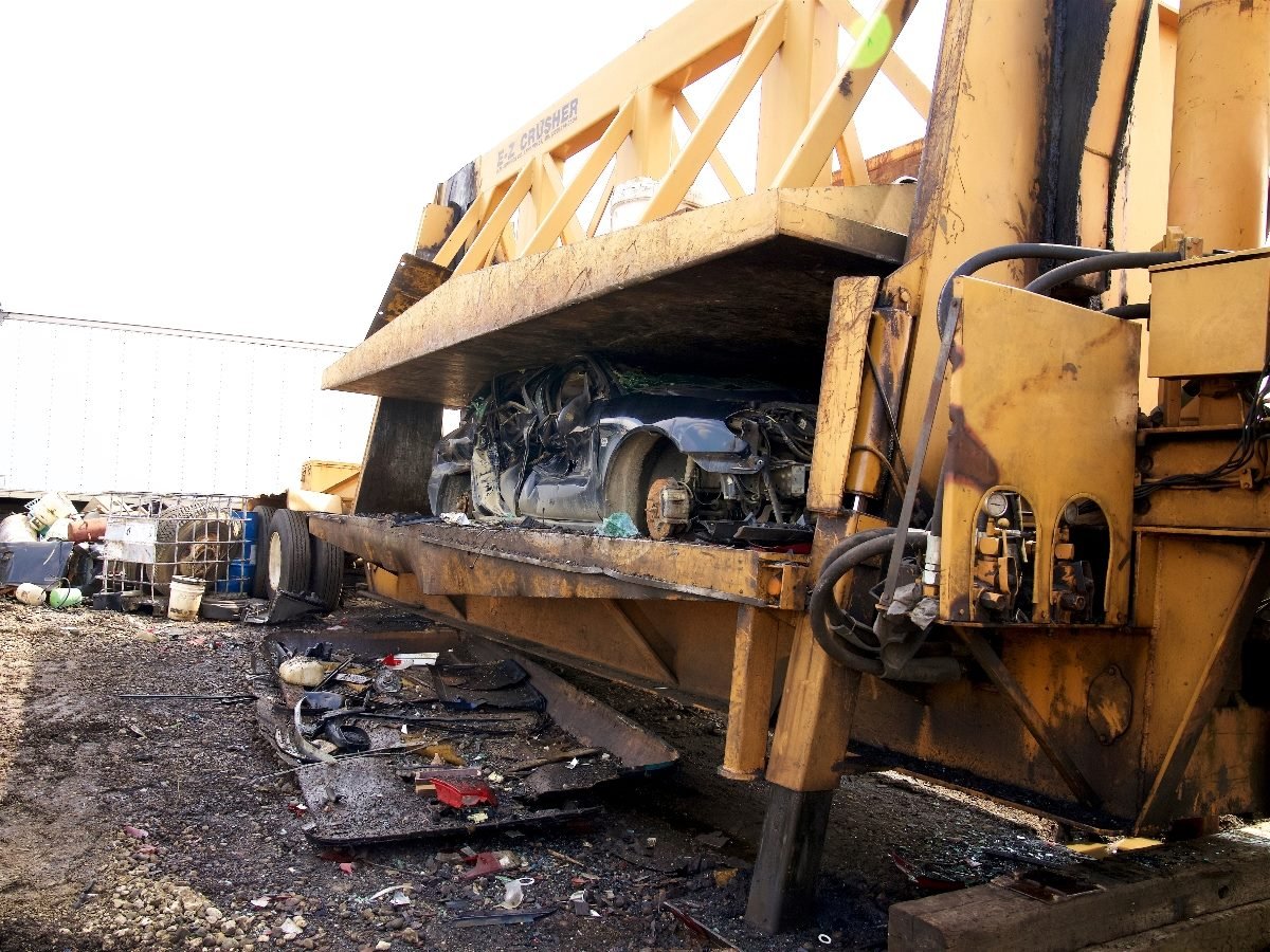 A large industrial car crusher compresses a vehicle into a flattened block of metal at a scrapyard, with scattered debris and machinery parts on the ground nearby.