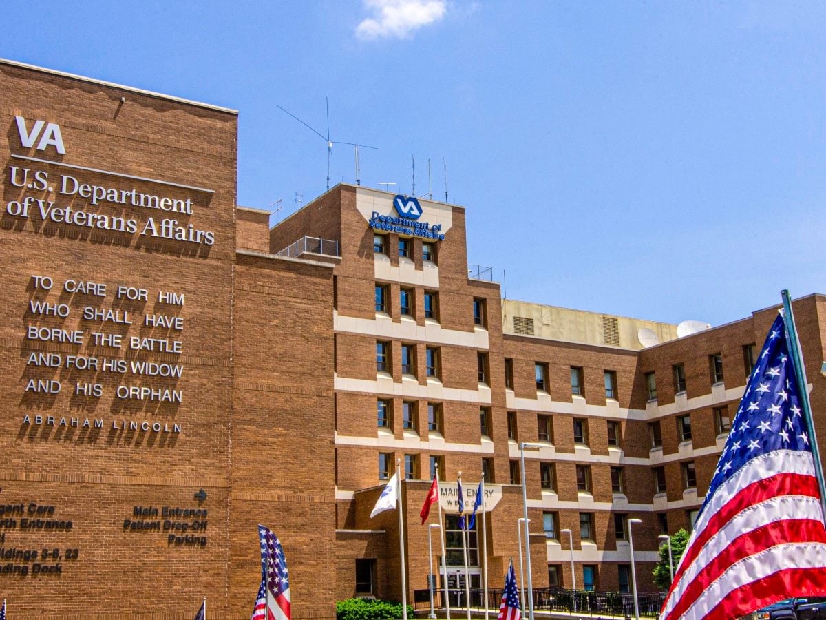 Exterior of a U.S. Department of Veterans Affairs medical center building with the VA logo, an Abraham Lincoln quote reading "To care for him who shall have borne the battle and for his widow and his orphan" engraved on the brick facade, and American flags displayed in front of the main entrance.