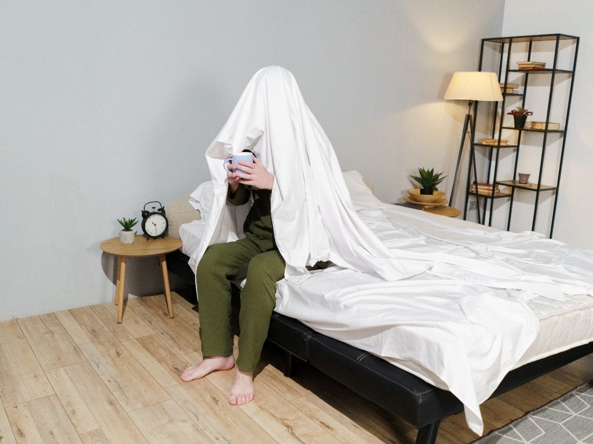 A person sitting on the edge of a bed with a white bedsheet draped over their head and shoulders, holding a smartphone in a softly lit bedroom with minimalist furniture.