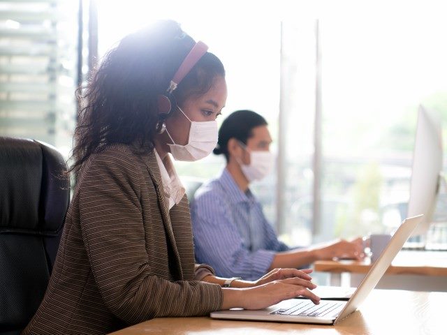 woman wearing a virus-prevention mask working on a laptop with a man in the background.