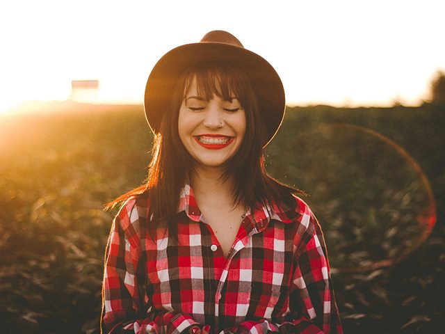 Woman with brown hair smiling with her eyes closed while in a field as the sun sets. 