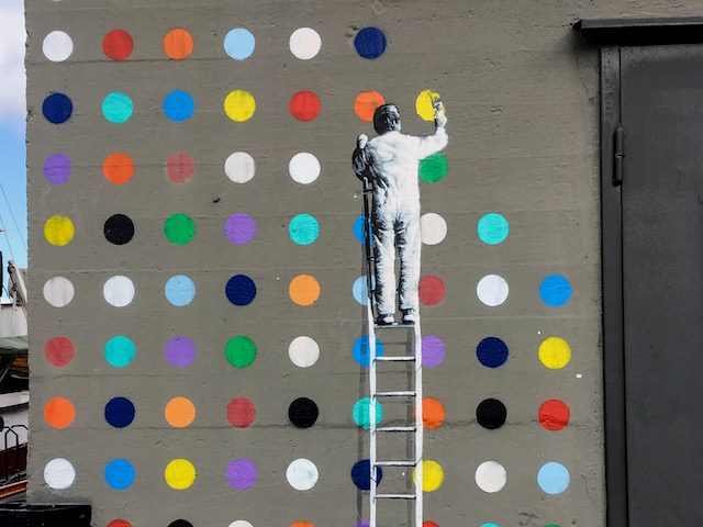 street art of a person standing on a ladder, painting a series of dots on a wall