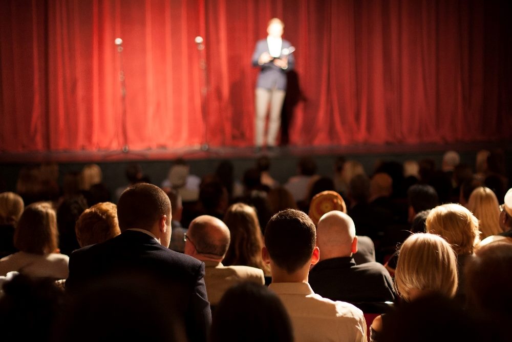 A comedian performs on a stage in front of a red curtain in front of a crowd in piece about EX and CX.