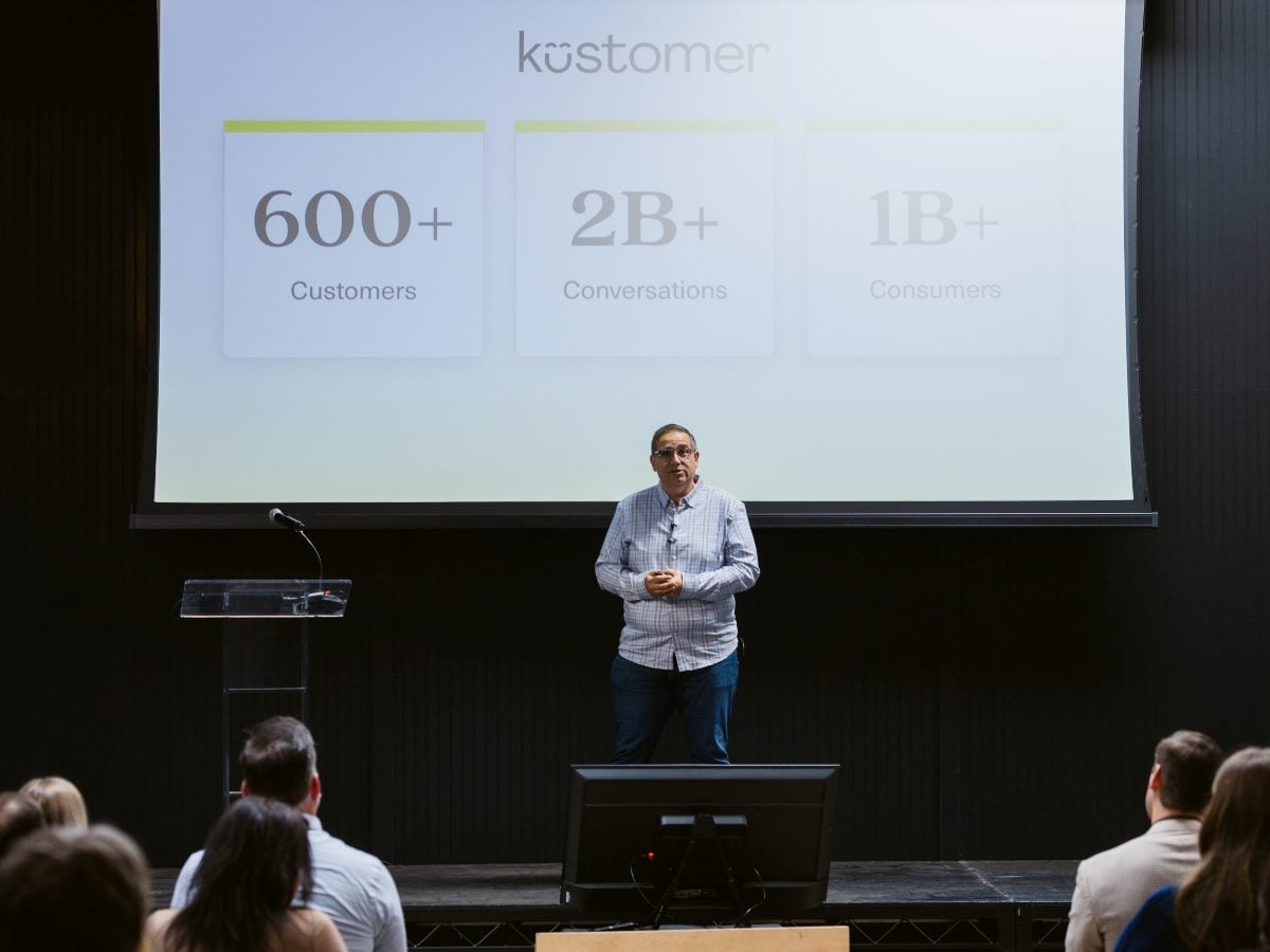 Brad Birnbaum, CEO and co-founder of Kustomer, stands on stage delivering a presentation to an audience, with a large screen behind him displaying customer and conversation metrics during a business event or conference session.