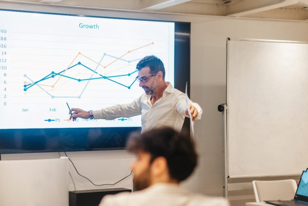 Business leader wearing glasses and holding a pen and piece of paper discusses a growth report projected on a screen behind them in a piece about data-driven decision-making.