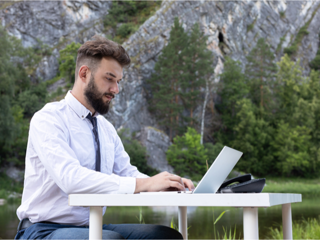 A businessman sitting at a desk working at his computer, situated in an out door environment with a mountainous background - Officeless workplace concept