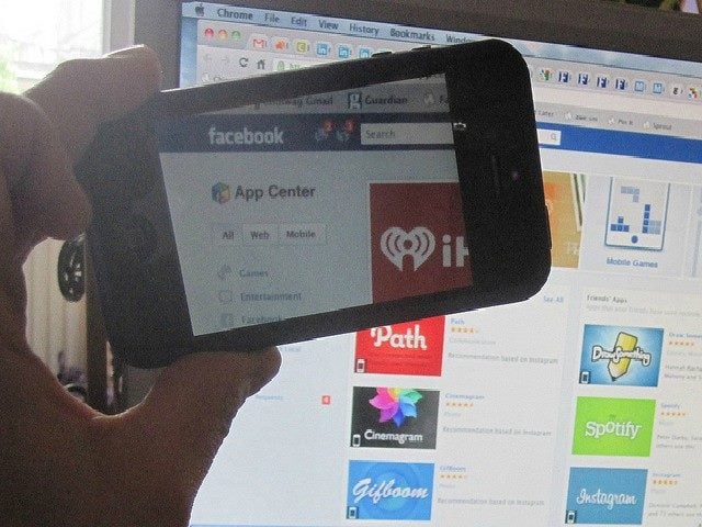 Someone holding a smart phone up to a computer screen taking a picture of a page with Facebook apps.