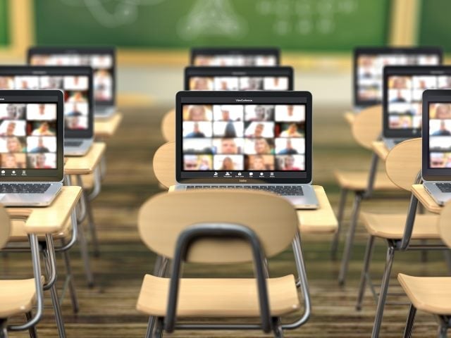 School desk and laptops with classroom app on the screen and blurred blackboard in the background. 