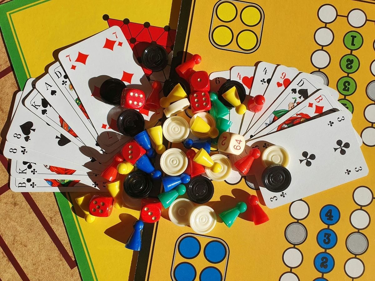 An overhead view of a colorful assortment of board game pieces, dice, checkers and playing cards scattered across a classic game board. Bright red, blue, green and yellow pawns are mixed with black and white checkers and fanned-out playing cards, creating a lively image that represents games, strategy, competition and chance.