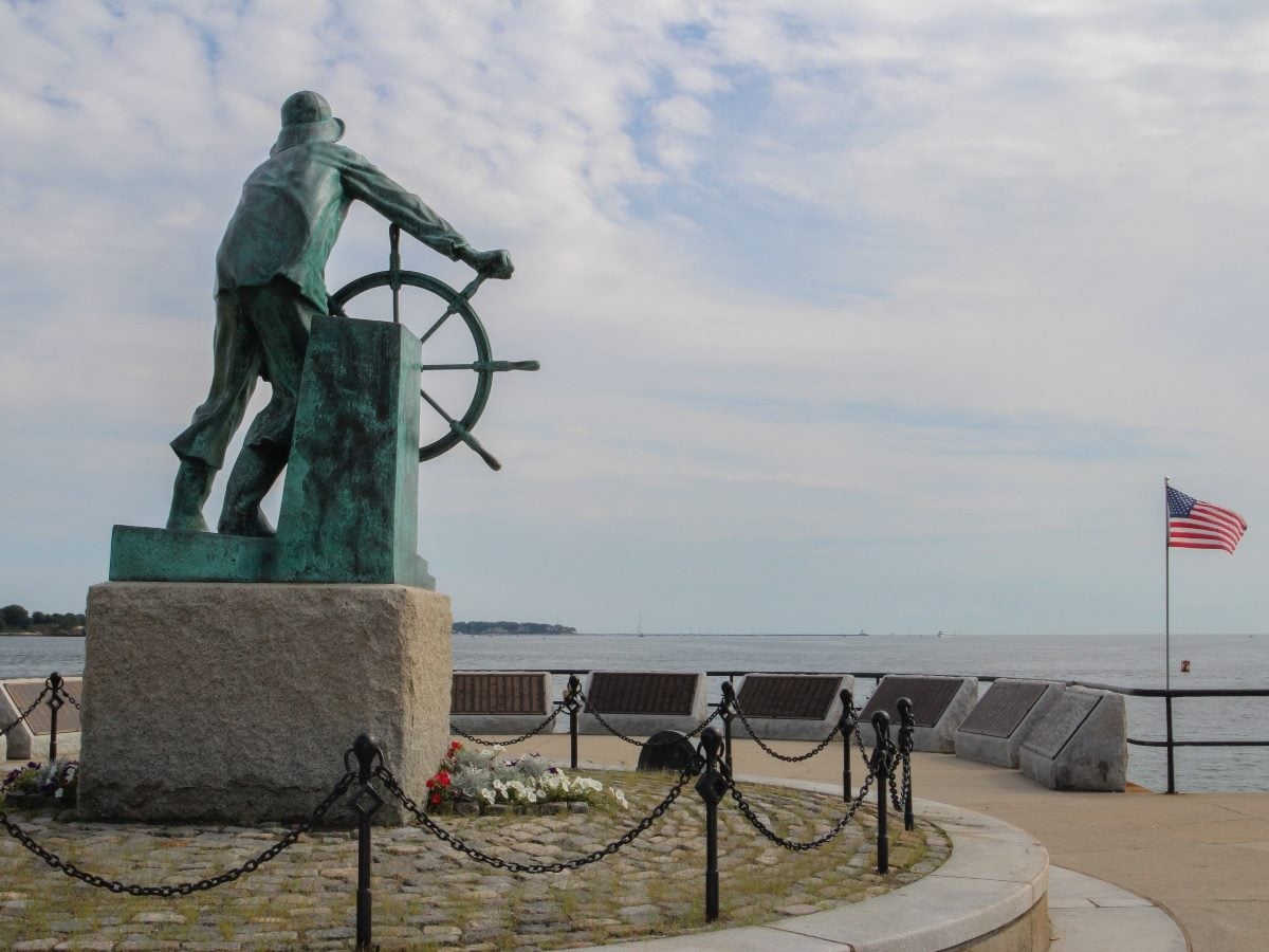 The Man at the Wheel statue, also known as the Gloucester Fisherman’s Memorial, stands on a granite pedestal overlooking the harbor in Gloucester, Massachusetts, with an American flag flying nearby.