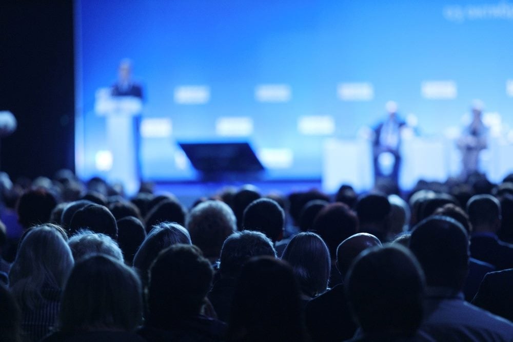 A speaker gives a talk in front of an audience in a conference hall lit in blue lighting in piece about contact center conferences and events.