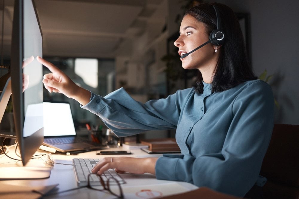 Call center, customer service employee at night working in front of a computer screen and pointing at computer screen while explaining something to a customer in piece about AI customer support.