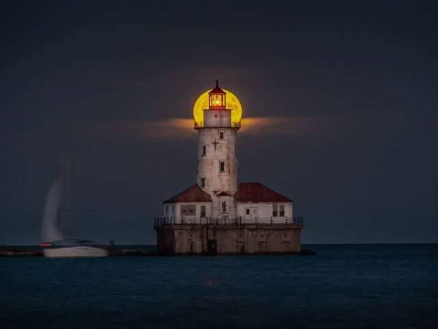 A beautiful night shot of the harvest full moon as it aligns with the center top of the abandoned historic light house tower along Lake Michigan in Chicago as sailboat passes by on the dark - Unified Insights concept