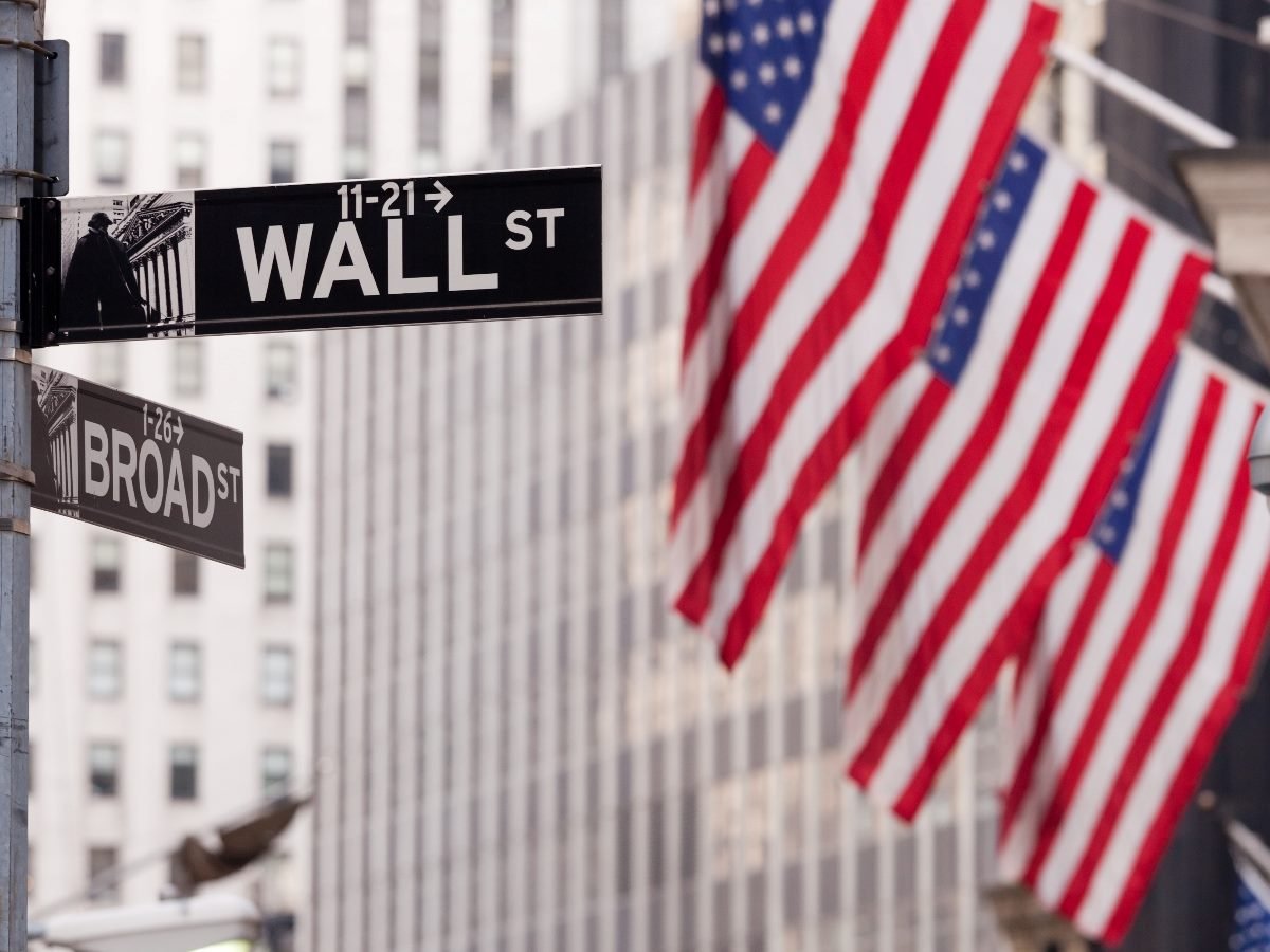 Street signs for Wall Street and Broad Street in New York’s Financial District, with multiple American flags hanging from nearby buildings in the background.