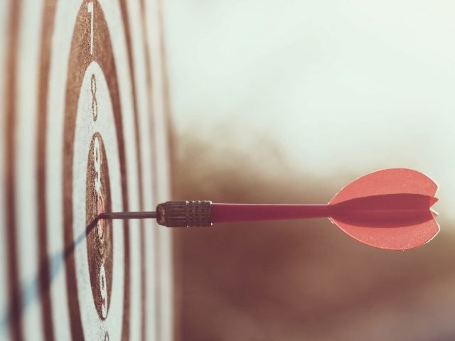 Close up red dart arrow hitting target center dartboard on sunset background.  