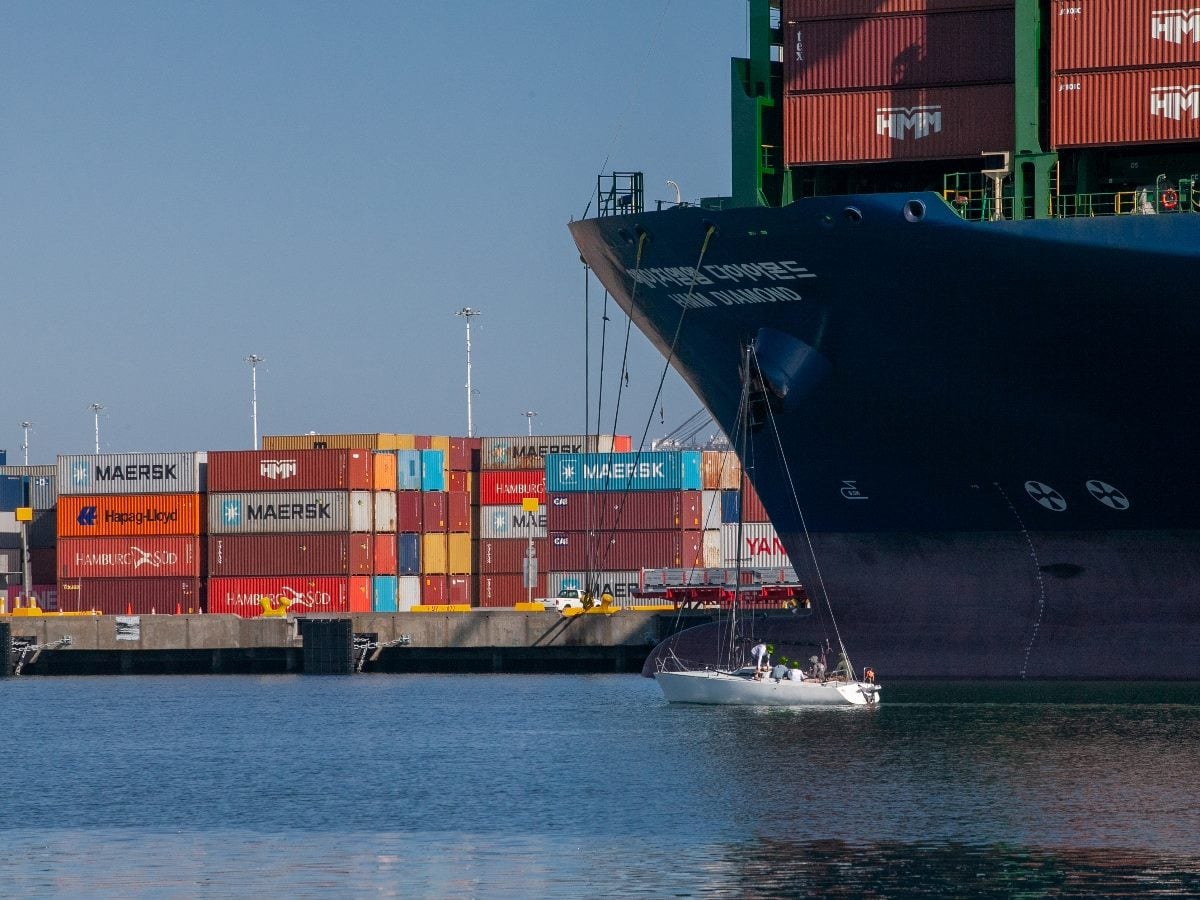 A Maersk container ship docked at a port with stacked shipping containers in the background, while a small sailboat passes in the foreground, highlighting the contrast between global logistics operations and individual maritime activity.