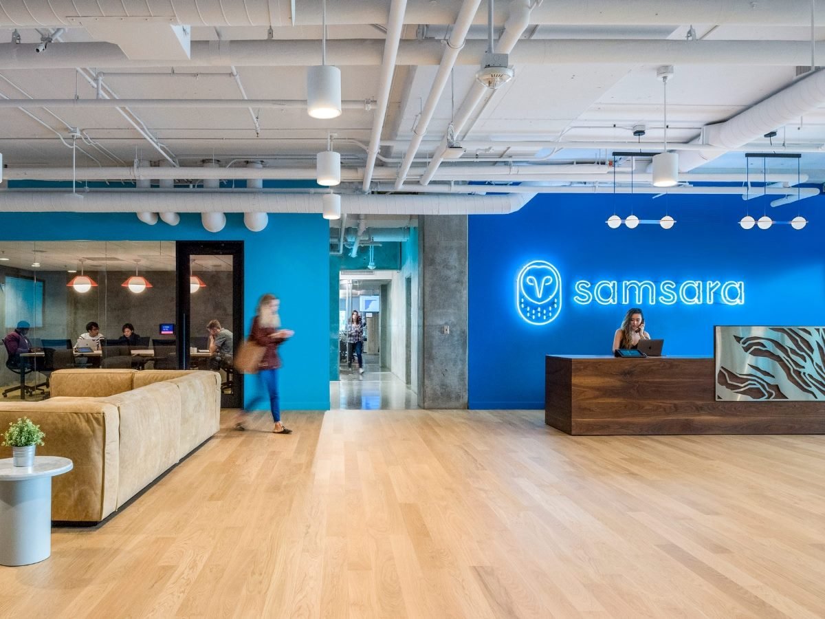 A modern office lobby with light wood floors and blue accent walls featuring a neon Samsara logo behind a wooden reception desk. A receptionist works at a laptop while a person walks by and several employees meet in a glass-walled conference room in the background.
