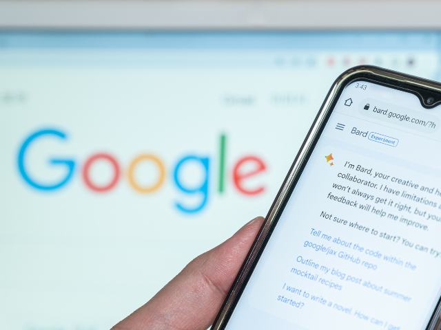A hand holding up a smartphone with Google Bard on the screen and the Google logo in the background 