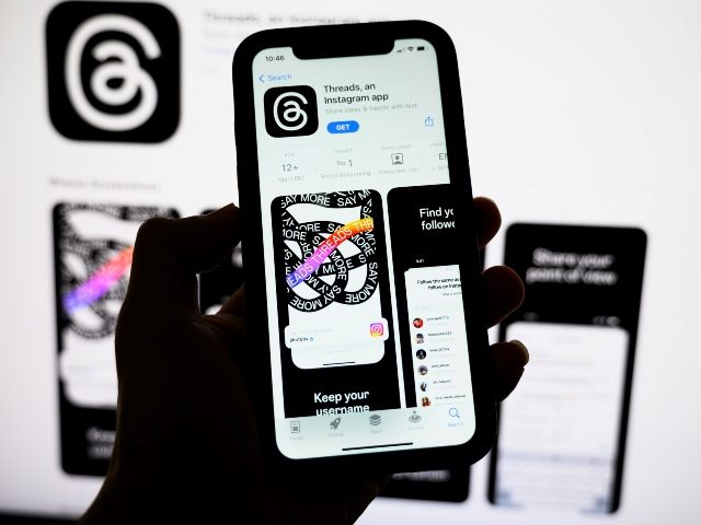 A hand holds a phone with Meta's Threads App on the screen and the black and white logo visible on a computer screen behind it, representing the possibility of advertising on Meta's  new Thread's social media platform. 