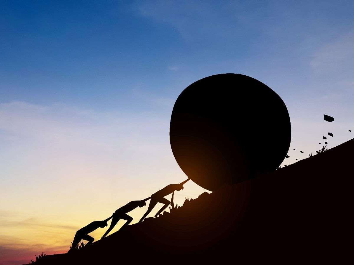 Silhouette illustration of several people pushing a massive boulder uphill on a steep slope at sunset, symbolizing effort, persistence, teamwork, friction and the challenge of overcoming obstacles to achieve a goal.