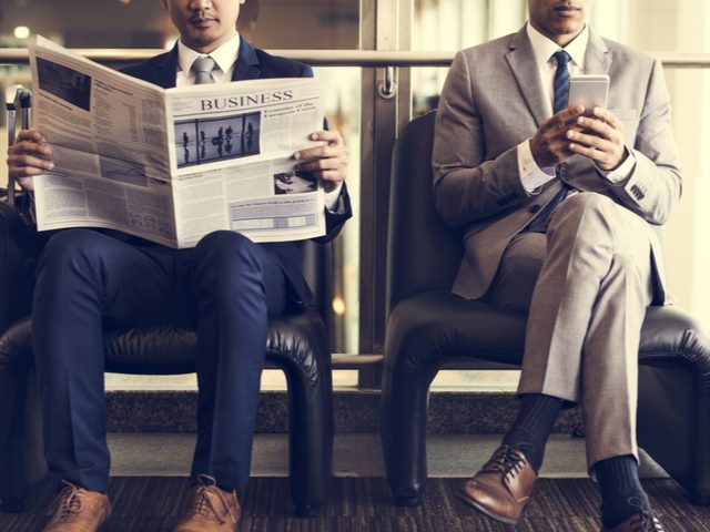 man in suit reading the newspaper next to a man reading on his mobile phone