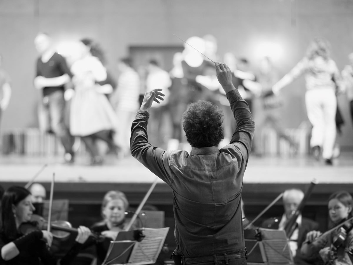 A conductor leads an orchestra during a rehearsal, with musicians playing string instruments in the foreground and dancers blurred in the background on stage.