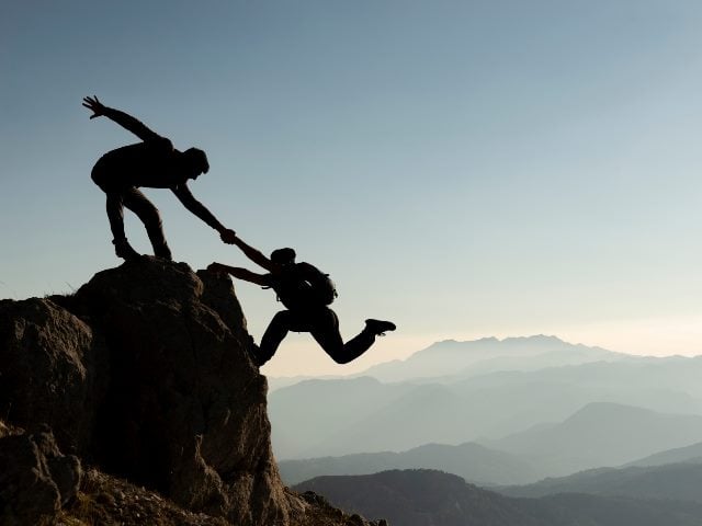 Silhouette image of one person on top of a mountain, grabbing the hand of another climber and lifting them up to the top. 