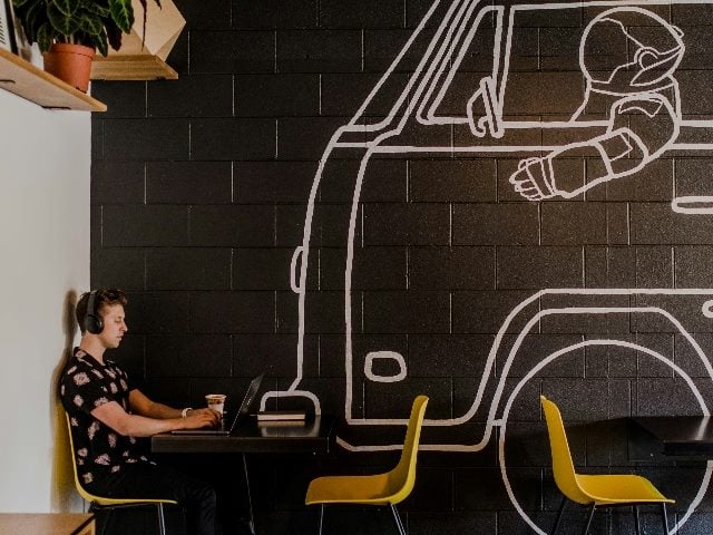 Man sitting at coffee shop laptop next to wall painted with robot driving a van.