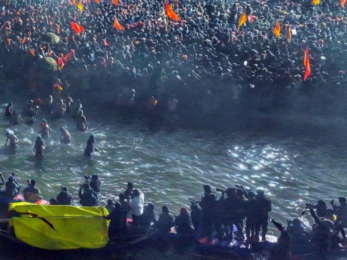 A massive crowd of devotees gathered along the riverbank at Maha Kumbh Mela, with worshippers bathing in the holy waters and saffron flags waving above the sea of people.