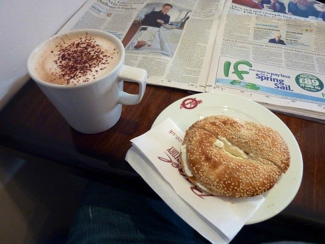 Bagel and cappuccino on a table next to a newspaper.