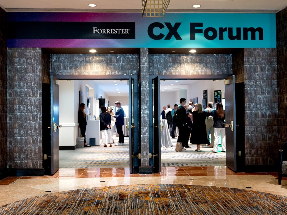 Entrance to the Forrester CX Forum event, featuring illuminated signage above open double doors leading into a conference networking space where attendees are gathered and conversing. The venue interior includes patterned carpeting, dark wood accents and event branding prominently displayed overhead.