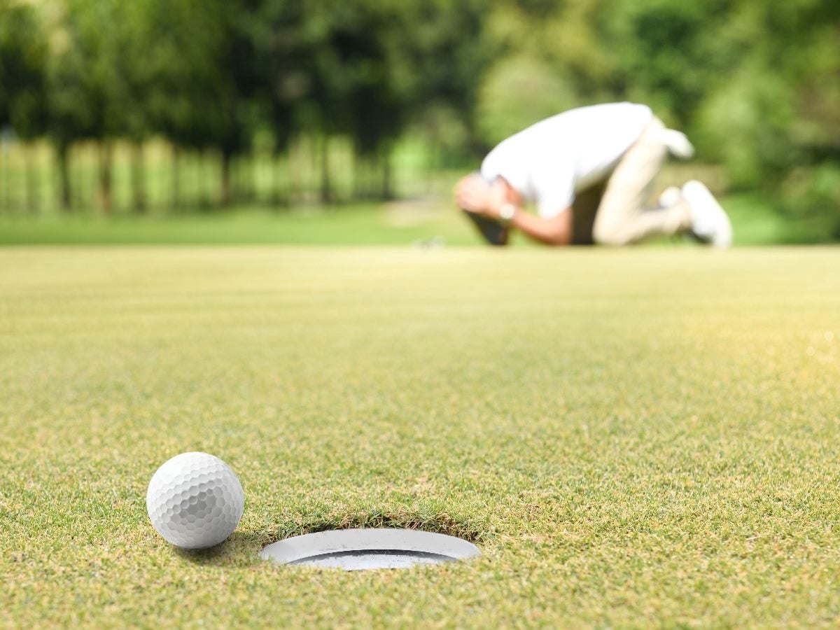 Golf ball resting inches from the hole on a putting green, with a blurred golfer kneeling in frustration in the background.