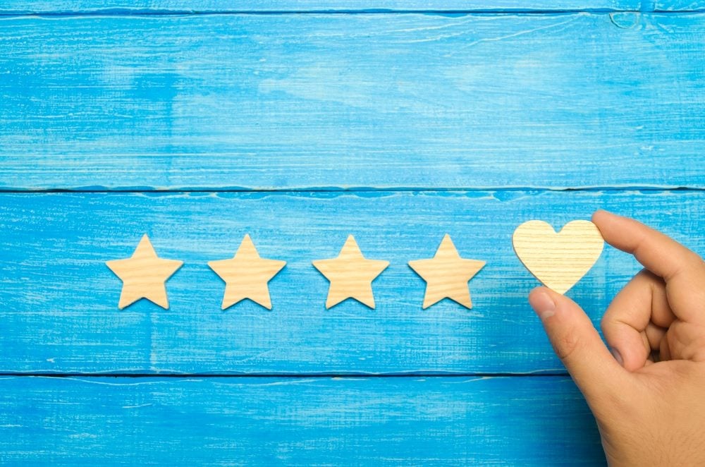 A hand places a golden ratings heart against 4 golden ratings stars against a bright blue wooden background in piece about contact centers and customer retention rates.