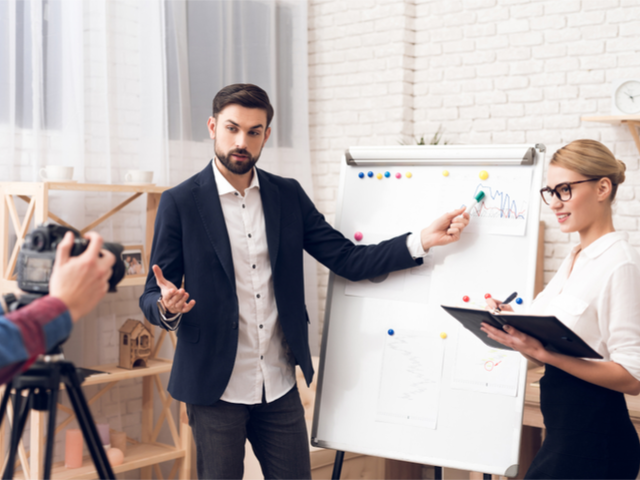 A marketing speaker in a suit and businesswoman in a blouse showing marketing charts at whiteboard while performing a marketing podcast