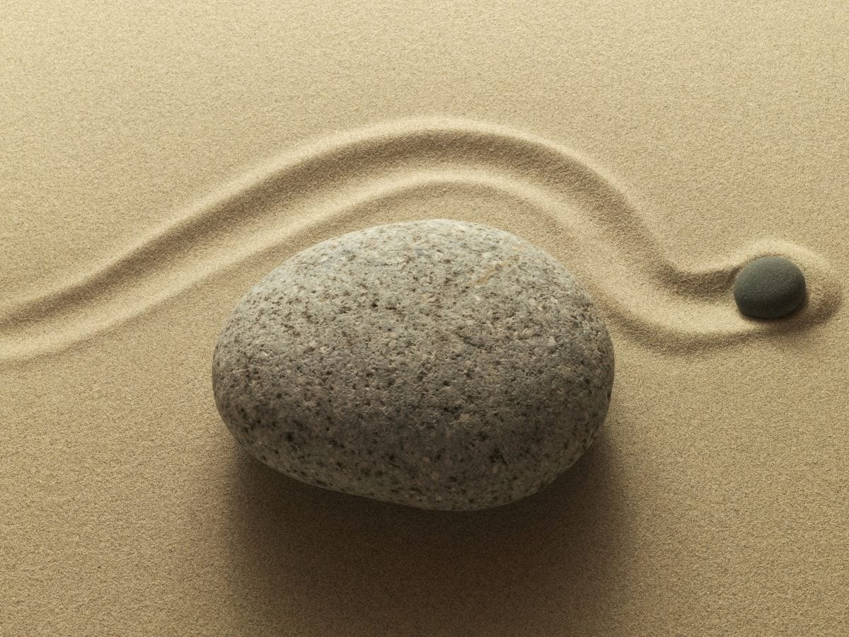 A smooth, rounded gray stone rests on finely raked beige sand, with a single curved groove flowing around the rock and leading to a smaller dark pebble nearby. The minimalist composition evokes a tranquil Zen garden with soft natural textures and balanced symmetry.