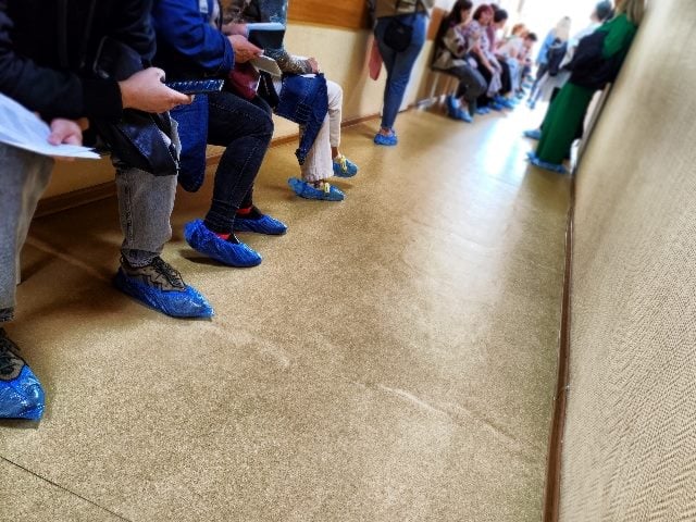 People sit in the hallway of a hospital waiting for a visit to the doctor. Shows peoples' legs as they sit down.