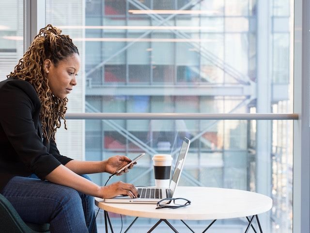 woman working remotely on a laptop and phone, plate glass window and high rise behind her