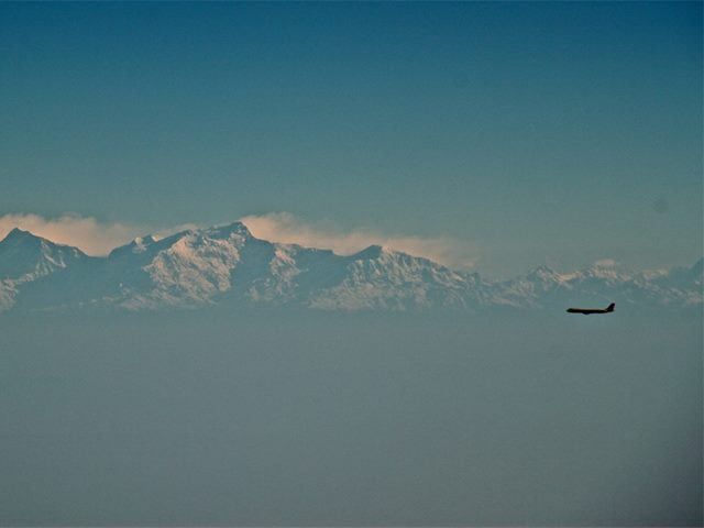 Image of large snow-capped mountains and a commercial airplane flying by, looking small in comparison to the mountains. 