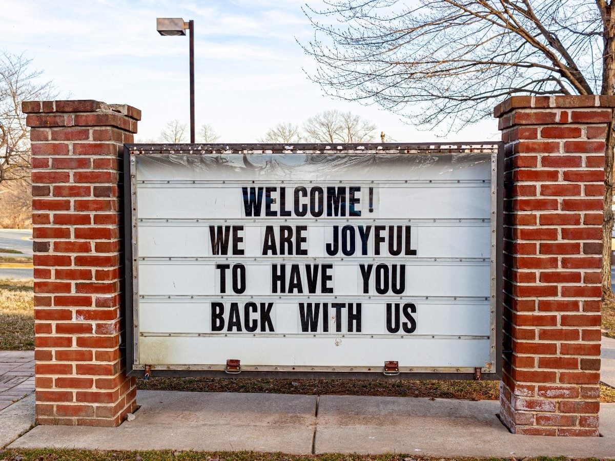 Outdoor marquee sign between two brick pillars reads, “WELCOME! WE ARE JOYFUL TO HAVE YOU BACK WITH US.”