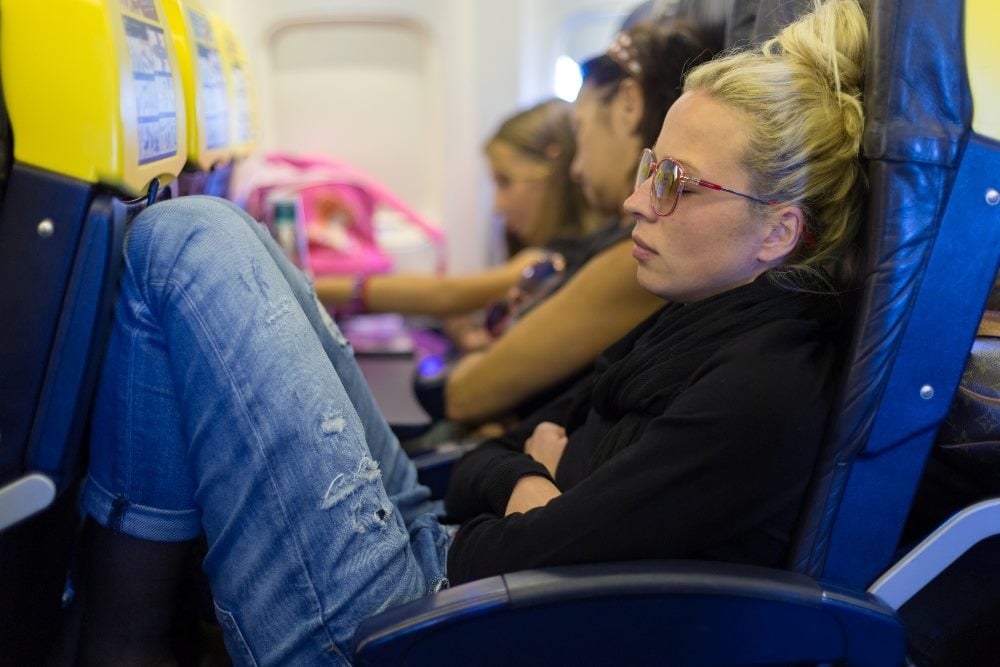 A row of tired airplane passengers with one person in the foreground wearing a jacket, old jeans and eye glasses sleeping in piece about improving customer experience the airline industry.