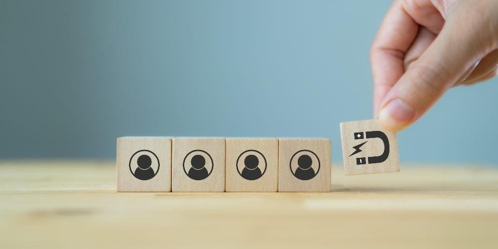A hand holds a wooden cube with a magnet emblazoned on it and holds in in front of four other wooden cubes with customers icons on them suggesting the importance of customer retention in piece about the customer retention rate.