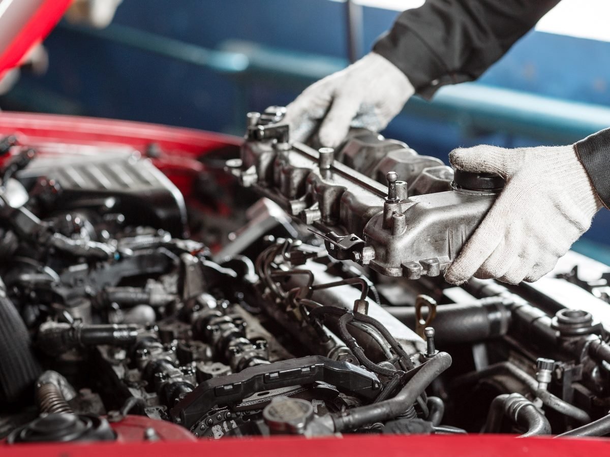 Mechanic wearing gloves removing a valve cover from a car engine during maintenance in a workshop.