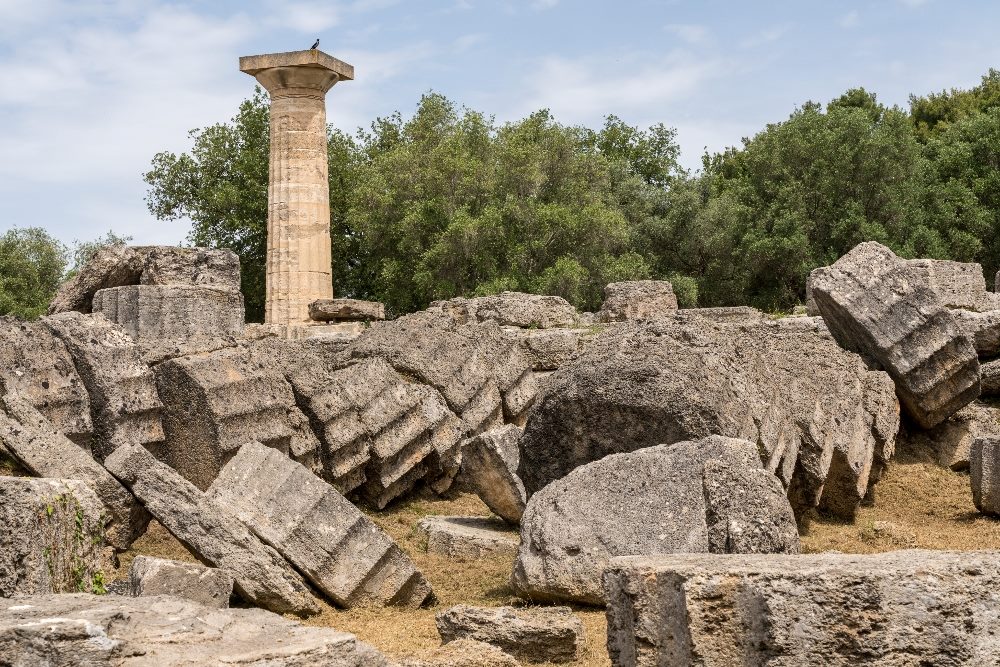Ruins of the Temple of Zeus at Olympia, Greece, symbolizing decay and collapse, akin to the decline in customer experience quality described in the Forrester report on CX decline. Scattered ancient stone blocks and a solitary column standing amid a lush green landscape evoke a poignant reminder of past grandeur and present deterioration.
