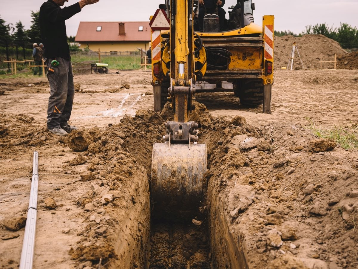 Construction worker directs a backhoe excavator as it digs a narrow trench through a dirt worksite. The excavator bucket is centered in the trench, with piles of excavated soil lining both sides and partially constructed residential buildings visible in the background.