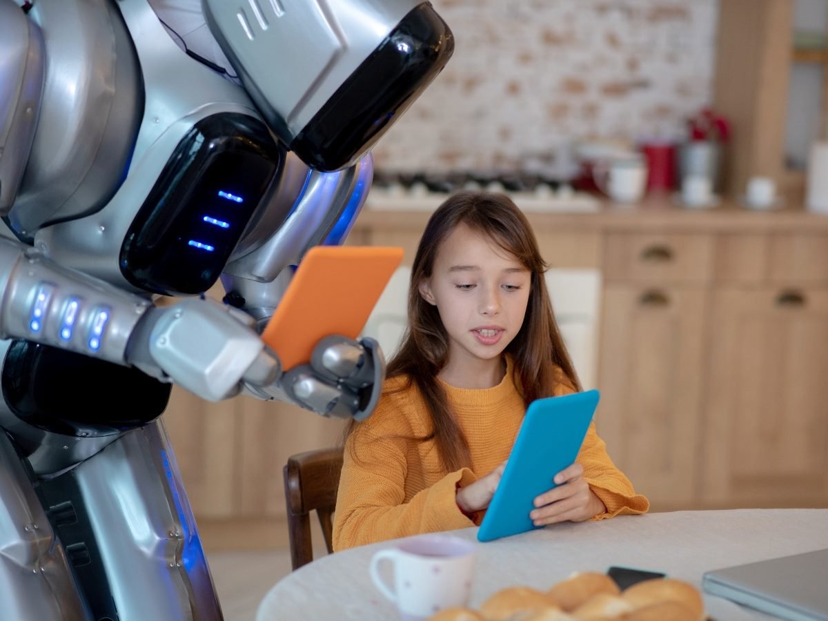 A humanoid robot standing beside a young girl at a kitchen table, both holding tablets as they look at their screens, suggesting a shared learning or technology interaction.