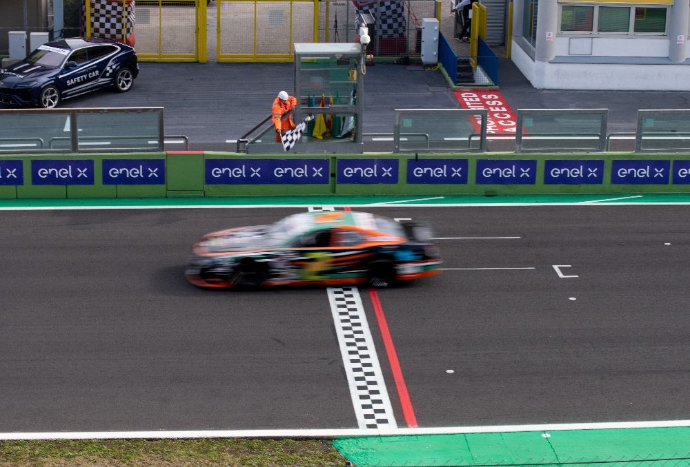 A race car blurs past the finish line, captured in motion on a race track. In the background, a safety car is parked beside the track, and a race official stands waving a checkered flag. The scene features prominent "Enel X" branding on the trackside barriers, suggesting sponsorship or partnership with the racing event. The track is bordered by a green curb and has various pit lane elements visible in piece about headless architecture.