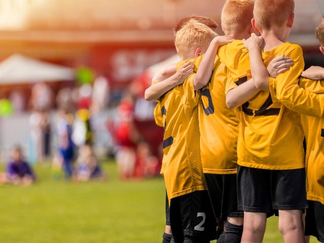 Kids sport team in yellow golden jerseys having pep talk with coach. Players have their arms around each other. 